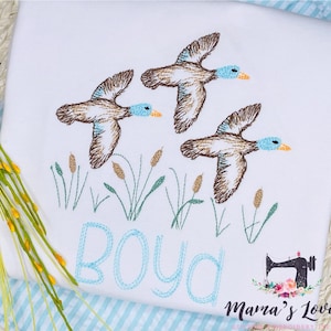 Puede incluir: Dise&ntilde;o bordado de tres patos marrones volando sobre juncos verdes con el nombre "Boyd" cosido en azul debajo.