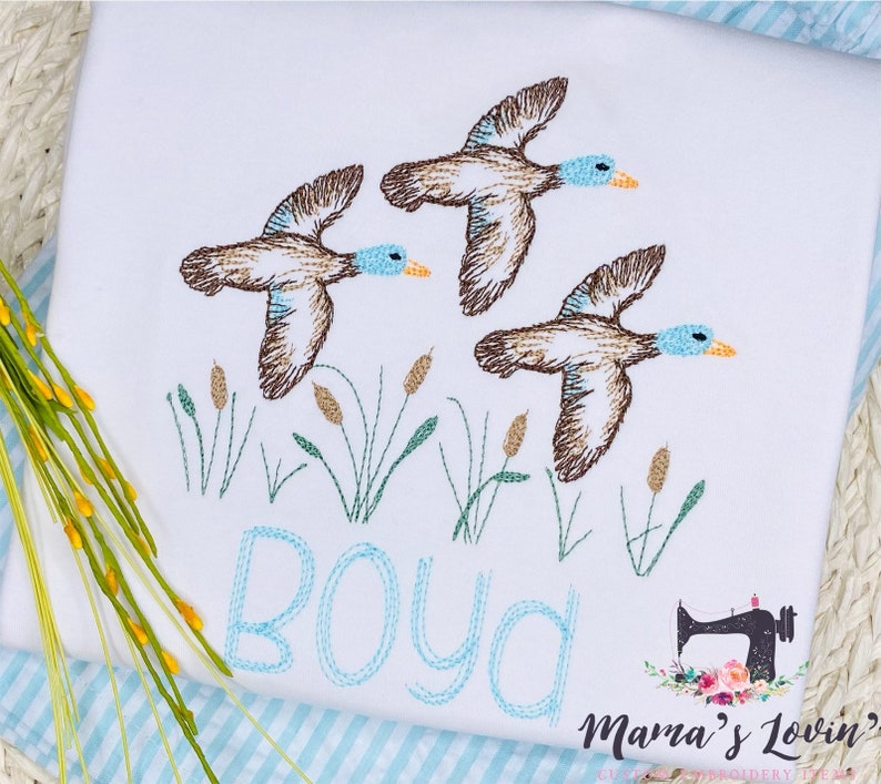 Puede incluir: Dise&ntilde;o bordado de tres patos marrones volando sobre juncos verdes con el nombre "Boyd" cosido en azul debajo.