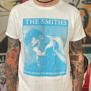 Puede incluir: Camiseta blanca con una imagen azul y blanca de un hombre sosteniendo un gato. El texto "THE SMITHS" está impreso en negro sobre la imagen y "HEAVEN KNOWS I'M MISERABLE MEOW" está impreso en negro debajo de la imagen.