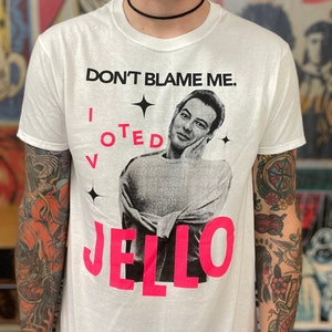 Peut inclure: T-shirt blanc avec un graphique rose d'un homme avec le texte "DON'T BLAME ME. I VOTED JELLO" imprimé dessus.