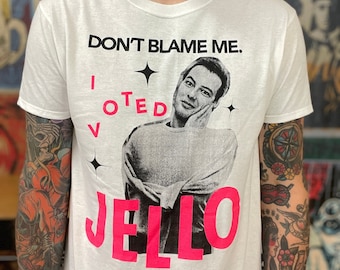 May include: A white short-sleeved t-shirt featuring a black and white graphic of a person with their hand on their cheek. The text reads DON'T BLAME ME. I VOTED JELLO in black and bright pink lettering, accented by four-pointed star symbols.