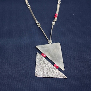 Puede incluir: Un collar de plata con un colgante geométrico que presenta dos triángulos superpuestos. El colgante está decorado con un patrón texturizado y tres pequeñas piedras preciosas rojas.