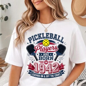May include: A white t-shirt featuring a graphic design with the text "PICKLEBALL Players ARE BORN IN 1942 LEGENDS ON & OFF THE COURT". The design includes a yellow pickleball, two dark blue paddles, and red and blue stars on a white background.