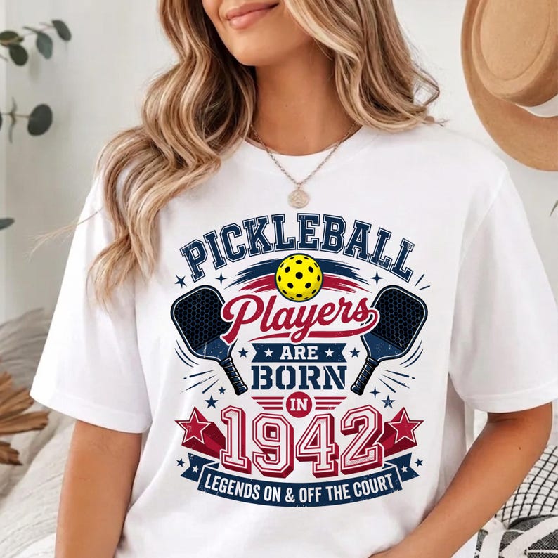May include: A white t-shirt featuring a graphic design with the text "PICKLEBALL Players ARE BORN IN 1942 LEGENDS ON & OFF THE COURT". The design includes a yellow pickleball, two dark blue paddles, and red and blue stars on a white background.