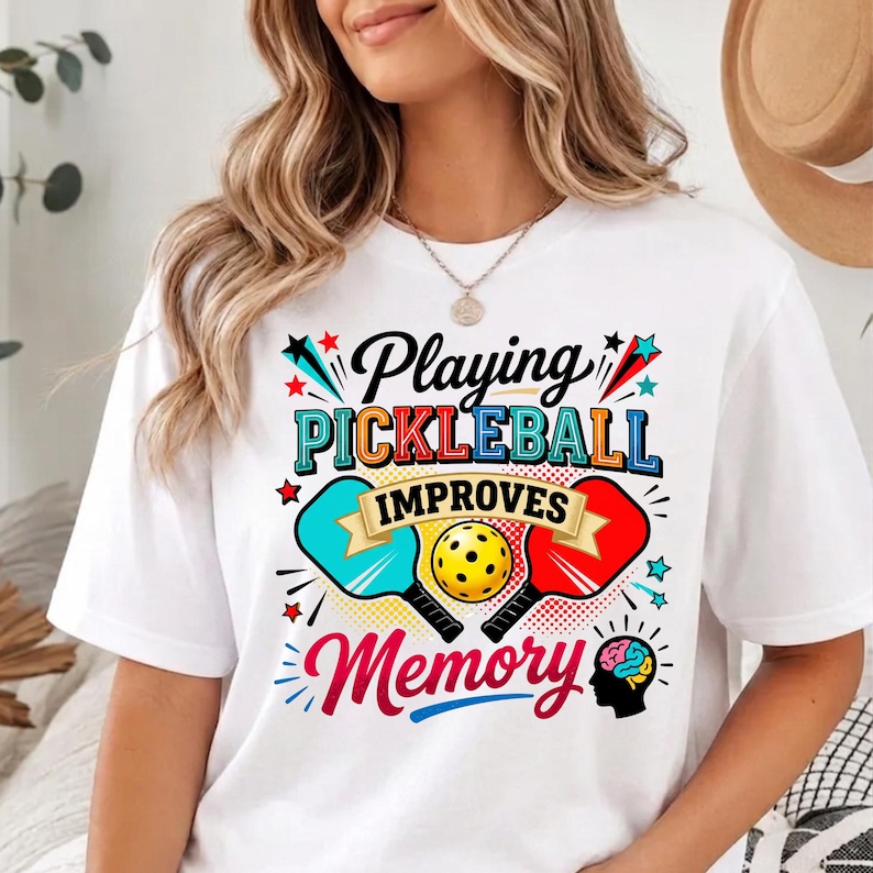 May include: A white t-shirt featuring a colorful graphic with the text Playing PICKLEBALL IMPROVES Memory. The design includes a yellow pickleball, a blue paddle, a red paddle, and a silhouette of a human head with a brain illustration.