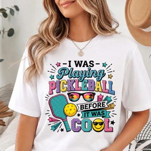 May include: A white t-shirt featuring a colorful graphic with the text "I WAS Playing PICKLEBALL BEFORE IT WAS COOL". The design includes a pickleball paddle, a yellow ball, sunglasses with a sunset reflection, and a smiling face with sunglasses.
