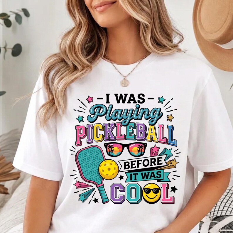 May include: A white t-shirt featuring a colorful graphic with the text "I WAS Playing PICKLEBALL BEFORE IT WAS COOL". The design includes a pickleball paddle, a yellow ball, sunglasses with a sunset reflection, and a smiling face with sunglasses.