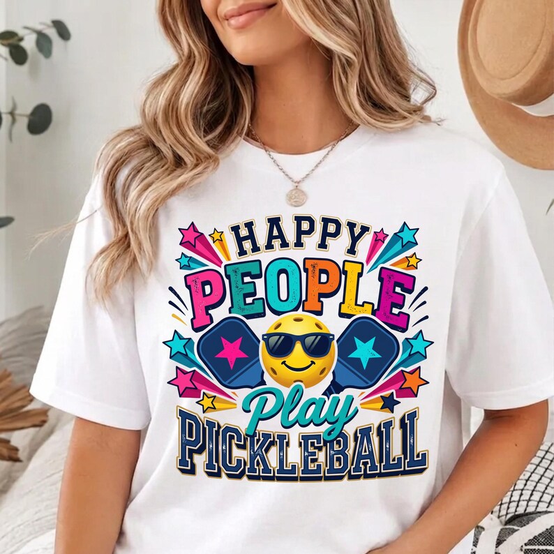May include: A white t-shirt featuring a colorful graphic with the text "HAPPY PEOPLE Play PICKLEBALL". The design includes a yellow smiley face wearing black sunglasses, two blue pickleball paddles with pink and blue stars, and various colorful starburst accents.