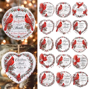 Può includere: Una collezione di ornamenti in ceramica rotondi e a forma di cuore con cardinali rossi, rami di pino e bacche di agrifoglio. Ogni ornamento mostra un testo nero in corsivo con temi commemorativi e festivi, incluse frasi come In Loving Memory e Christmas Leaves a Heart.