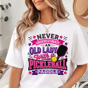 May include: A white short-sleeve t-shirt featuring a graphic design with the text "NEVER UNDERESTIMATE AN OLD LADY With a PICKLEBALL PADDLE". The design includes a black pickleball paddle, a yellow pickleball, and pink and purple stars.