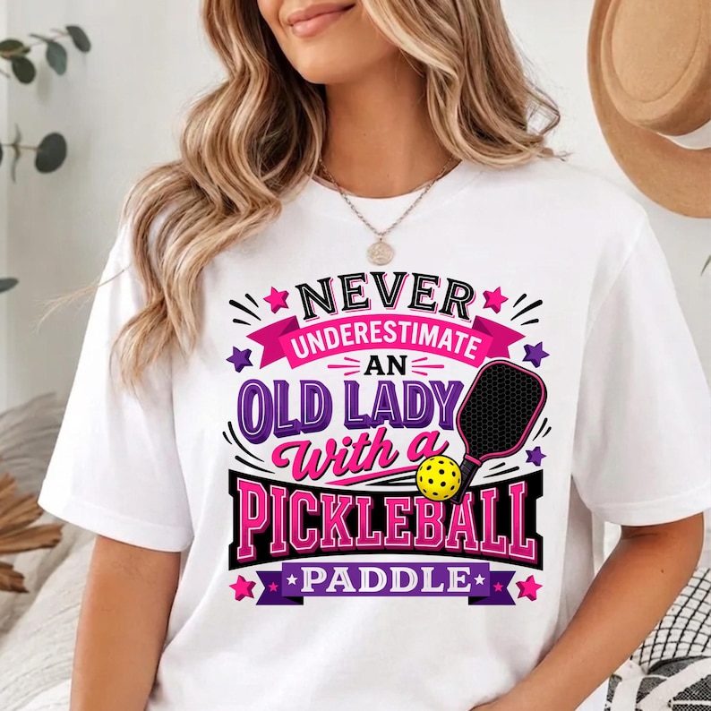May include: A white short-sleeve t-shirt featuring a graphic design with the text "NEVER UNDERESTIMATE AN OLD LADY With a PICKLEBALL PADDLE". The design includes a black pickleball paddle, a yellow pickleball, and pink and purple stars.