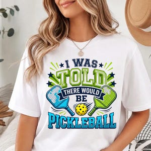 May include: A white short-sleeved t-shirt featuring a graphic print with the text I WAS TOLD THERE WOULD BE PICKLEBALL. The design includes two pickleball paddles in blue and green, a yellow pickleball, and dark blue star accents.