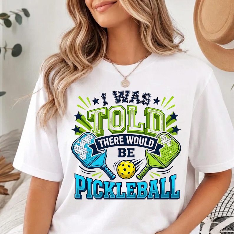 May include: A white short-sleeved t-shirt featuring a graphic print with the text I WAS TOLD THERE WOULD BE PICKLEBALL. The design includes two pickleball paddles in blue and green, a yellow pickleball, and dark blue star accents.