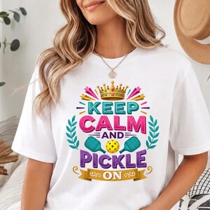 May include: A white short-sleeved t-shirt featuring a colorful graphic print that reads KEEP CALM AND PICKLE ON. The design includes a gold crown, two teal pickleball paddles, a yellow pickleball, and decorative teal leaf accents.