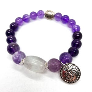 May include: Purple amethyst gemstone bracelet with a silver charm featuring the Om symbol. The bracelet has a gradient of purple shades and a clear crystal bead.