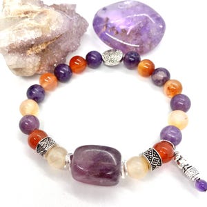 May include: A beaded bracelet featuring a variety of gemstones in shades of purple, orange, and cream. The bracelet includes a large, rectangular purple stone and decorative silver-colored metal accents. Additional purple and cream-colored stones are visible in the background.