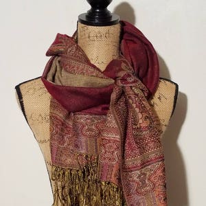Puede incluir: Una bufanda de color burdeos y dorado con estampado paisley y flecos. La bufanda está drapeada alrededor del cuello de un maniquí.