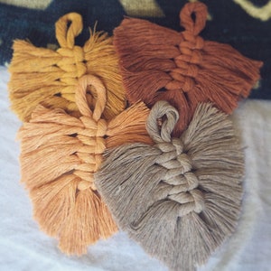 Puede incluir: Cuatro colgantes de pared de macram&eacute; hechos a mano en forma de hoja, en diferentes tonos de marr&oacute;n, naranja y amarillo. Las hojas est&aacute;n hechas con un cord&oacute;n grueso de fibra natural y tienen un borde con flecos.