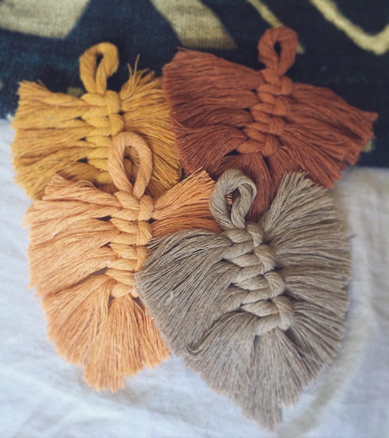 Puede incluir: Cuatro colgantes de pared de macram&eacute; hechos a mano en forma de hoja, en diferentes tonos de marr&oacute;n, naranja y amarillo. Las hojas est&aacute;n hechas con un cord&oacute;n grueso de fibra natural y tienen un borde con flecos.