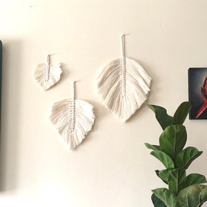 May include: Three white macrame leaf wall hangings of varying sizes. The largest leaf is at the top, the smallest leaf is at the bottom, and the middle-sized leaf is in between. The leaves are hanging on a white wall.