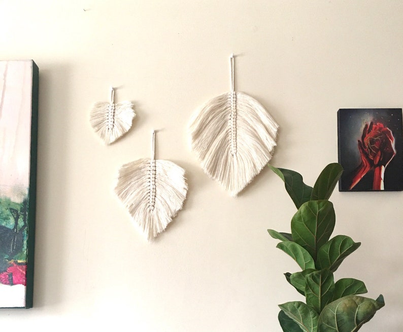 May include: Three white macrame leaf wall hangings of varying sizes. The largest leaf is at the top, the smallest leaf is at the bottom, and the middle-sized leaf is in between. The leaves are hanging on a white wall.