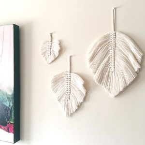 May include: Three white macrame feather wall hangings of varying sizes. The largest feather is at the top, the smallest is at the bottom, and the middle feather is in between.