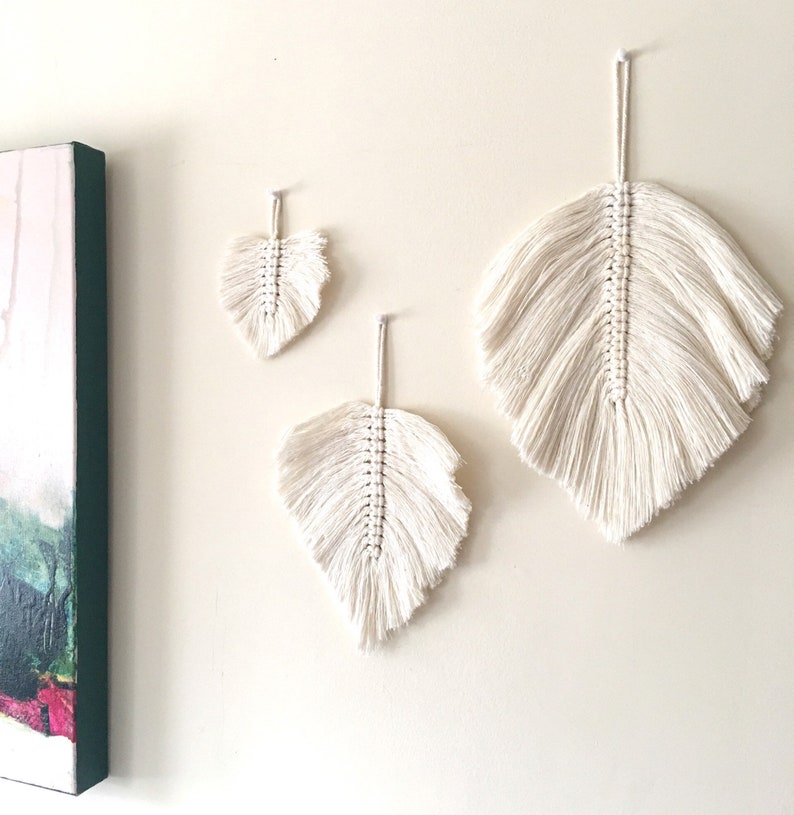 May include: Three white macrame feather wall hangings of varying sizes. The largest feather is at the top, the smallest is at the bottom, and the middle feather is in between.