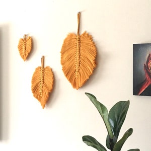 Puede incluir: Tres tapices de macram&eacute; en forma de hojas, hechos con hilo amarillo mostaza. Las hojas est&aacute;n colgadas en una pared blanca.