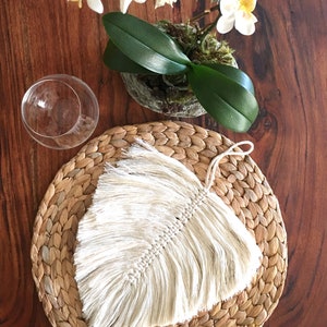 May include: A white macrame feather with a fringe hanging on a round woven straw placemat. The placemat is on a wooden table.