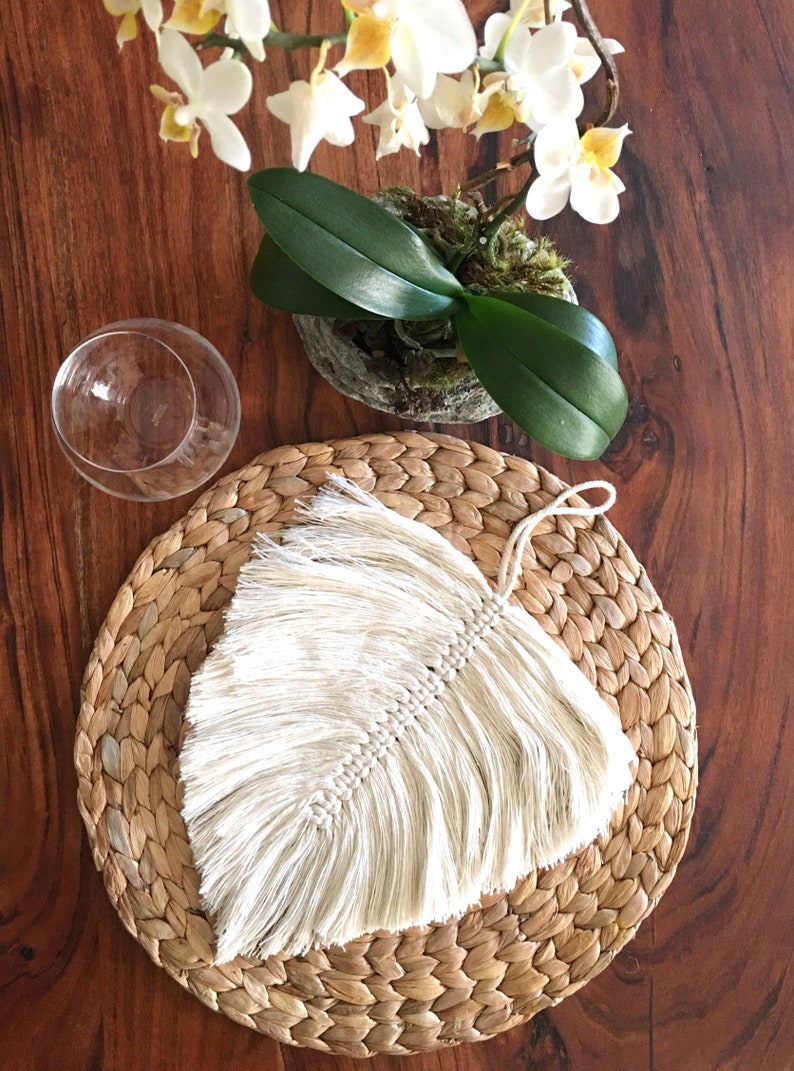 May include: A white macrame feather with a fringe hanging on a round woven straw placemat. The placemat is on a wooden table.
