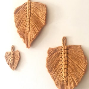 Puede incluir: Tres tapices de macram&eacute; en forma de hojas, hechos con cord&oacute;n de algod&oacute;n beige natural. Las hojas son de diferentes tama&ntilde;os, con la hoja m&aacute;s grande en la parte superior y la hoja m&aacute;s peque&ntilde;a en la parte inferior.