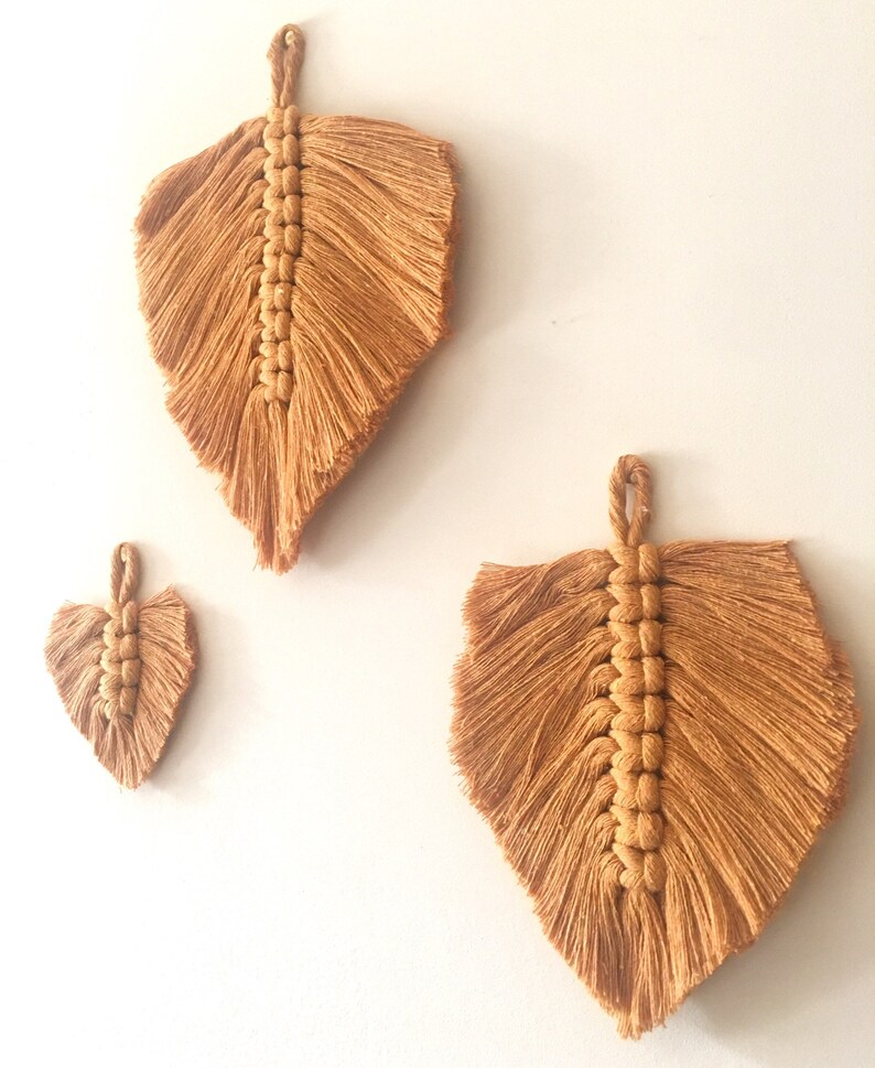 Puede incluir: Tres tapices de macram&eacute; en forma de hojas, hechos con cord&oacute;n de algod&oacute;n beige natural. Las hojas son de diferentes tama&ntilde;os, con la hoja m&aacute;s grande en la parte superior y la hoja m&aacute;s peque&ntilde;a en la parte inferior.