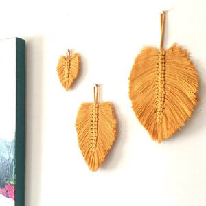 Puede incluir: Tres colgantes de pared de plumas de macram&eacute; de color amarillo en diferentes tama&ntilde;os. La pluma m&aacute;s grande est&aacute; en la parte superior de la imagen, y la pluma m&aacute;s peque&ntilde;a est&aacute; en la parte inferior.