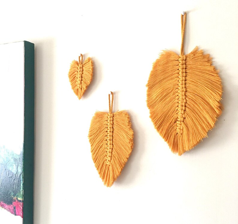 Puede incluir: Tres colgantes de pared de plumas de macram&eacute; de color amarillo en diferentes tama&ntilde;os. La pluma m&aacute;s grande est&aacute; en la parte superior de la imagen, y la pluma m&aacute;s peque&ntilde;a est&aacute; en la parte inferior.