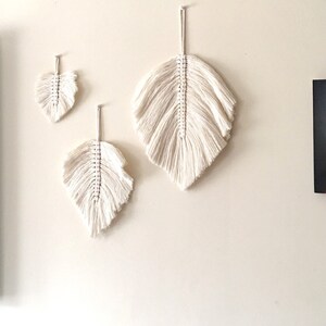 May include: Three white macrame feather wall hangings of varying sizes. The largest feather is at the top, and the smallest feather is at the bottom.
