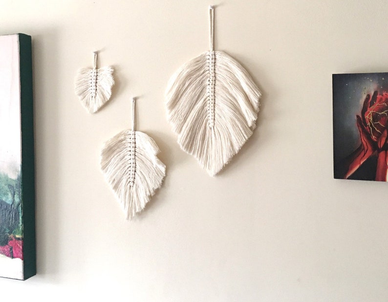 May include: Three white macrame feather wall hangings of varying sizes. The largest feather is at the top, and the smallest feather is at the bottom.