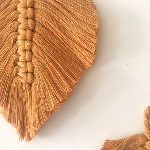 Puede incluir: Dos tapices de pared de plumas de macram&eacute; de color mostaza con detalles de flecos.
