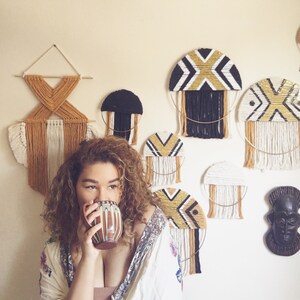 Puede incluir: Una mujer con el pelo rizado sostiene una taza de cer&aacute;mica marr&oacute;n y blanca. La pared detr&aacute;s de ella est&aacute; decorada con tapices de macram&eacute; en negro, blanco y dorado.