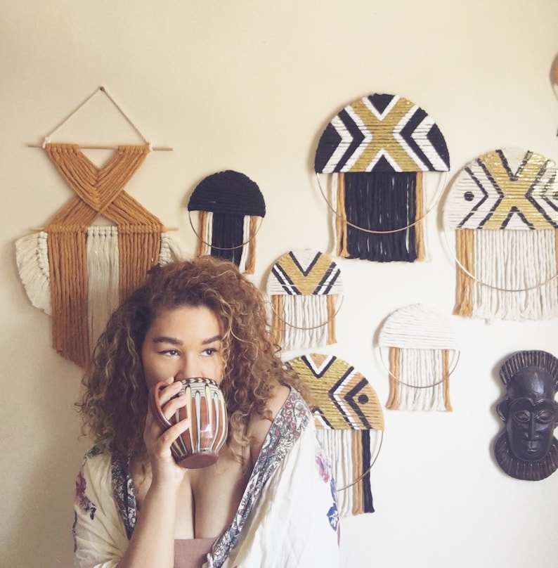 Puede incluir: Una mujer con el pelo rizado sostiene una taza de cer&aacute;mica marr&oacute;n y blanca. La pared detr&aacute;s de ella est&aacute; decorada con tapices de macram&eacute; en negro, blanco y dorado.