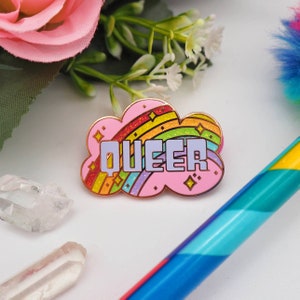 May include: A pink cloud-shaped enamel pin with a rainbow and glitter design. The word "QUEER" is written in purple letters on the pin.