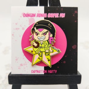 Op de afbeelding: Een gouden emaille pin met een cartoonfiguur met roze haar, een gele jumpsuit en een zwarte pet. Het personage zit op een gouden ster en houdt een bruin voorwerp vast. De pin is op een witte en zwarte achtergrond met de tekst "DANGAN RONPA ENAMEL PIN" en "DAMNATION PARTY".
