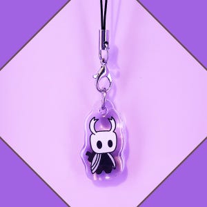 May include: A clear acrylic charm with a black and white illustration of the Knight character from the video game Hollow Knight. The charm is attached to a black lanyard with a silver clasp.