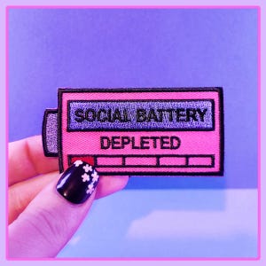 May include: A pink embroidered patch with the words "SOCIAL BATTERY" and "DEPLETED" in a rectangular design. The patch resembles a battery indicator, with a low charge level. The patch is held up by a hand with a black nail with white flower designs.