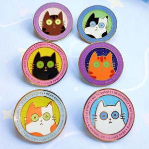Op de afbeelding: Vijf emaille-pins met verschillende katten met de tekst "Head Empty, No Thoughts" rond de rand van elke pin. De pins bevinden zich op een lichtblauwe achtergrond met witte sterren.