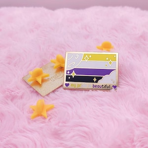 Op de afbeelding: Een gouden emaille pin met een gestileerde afbeelding van de non-binaire pride-vlag. De vlag heeft gele, witte, paarse en zwarte strepen met een halve maan en sterren. De pin heeft de tekst "my pride is beautiful" in gouden letters.