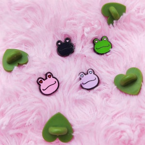 May include: Five enamel pins featuring frogs with hearts for eyes. The frogs are black, green, pink, and purple. The pins are on a pink fluffy background.