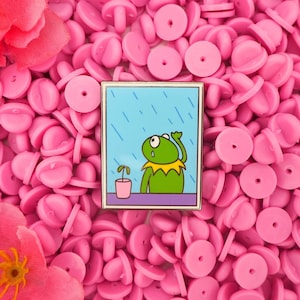 May include: A rectangular enamel pin depicting a cartoon frog looking sad in the rain. The frog is green with a yellow collar, and a small potted plant sits on a table. The background is a light blue with rain lines.