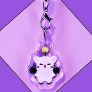 May include: A clear acrylic keychain featuring a white cat with black bat wings and a yellow star above it. The keychain is attached to a silver metal clasp.