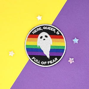 May include: A round rainbow-coloured patch with a white ghost in the centre. The text "Here, Queer, & Full of Fear" is written around the edge of the patch.