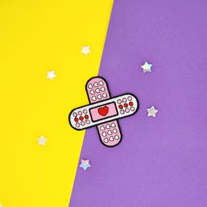 May include: Pink fabric patch shaped like a bandage with a red heart in the center. The patch is decorated with small white hearts.